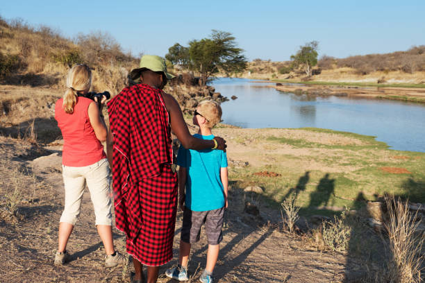 Ruaha National Park,Tanzania - August 07, 2018: Rear view of a Masai guide in traditional red clothing, standing near some tourists  at the edge of the river during a game drive  in the Ruaha National Park. They are looking for wildlife near the river in the afternoon.