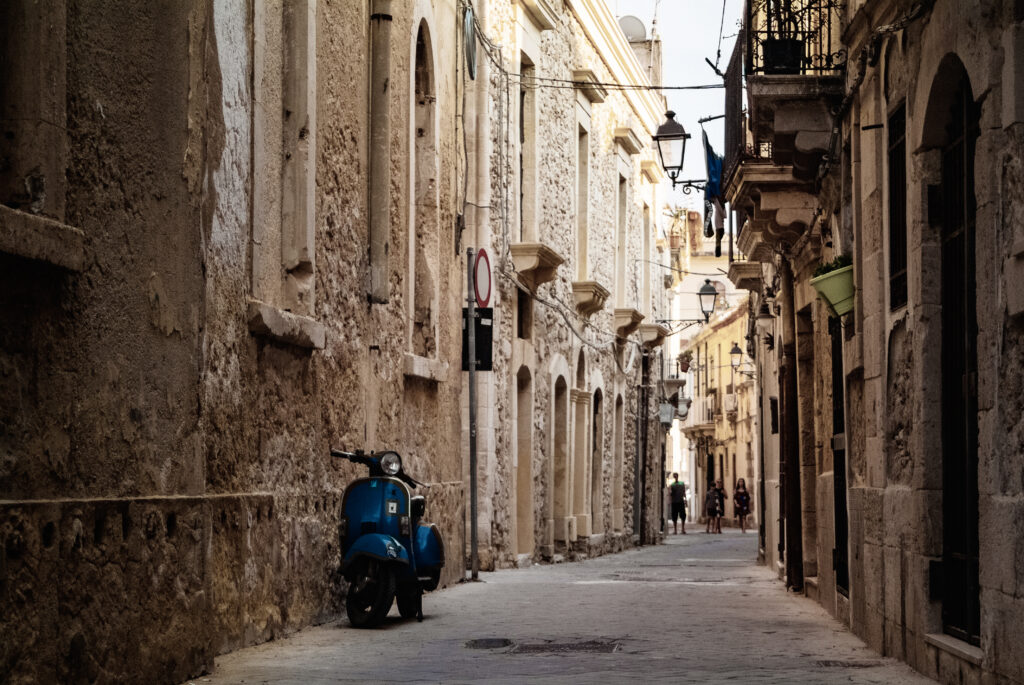 Blue Piaggio Vespa parked at sunset while people walking on the street. Ortigia urban area. Syracuse Siracusa, Sicily Italy, summer season