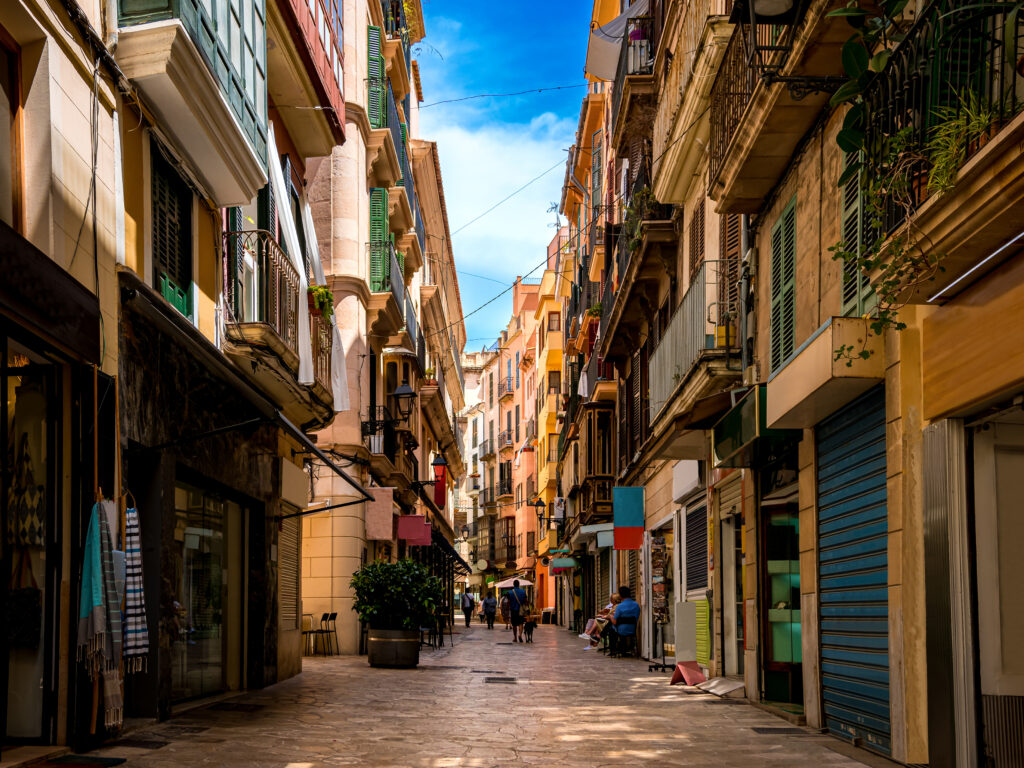 Few tourists walk through a narrow Mediterranean alley with small shops and restaurants in the shadow of residential buildings in the groomed center of Palma de Mallorca on an idyllic day in summer.