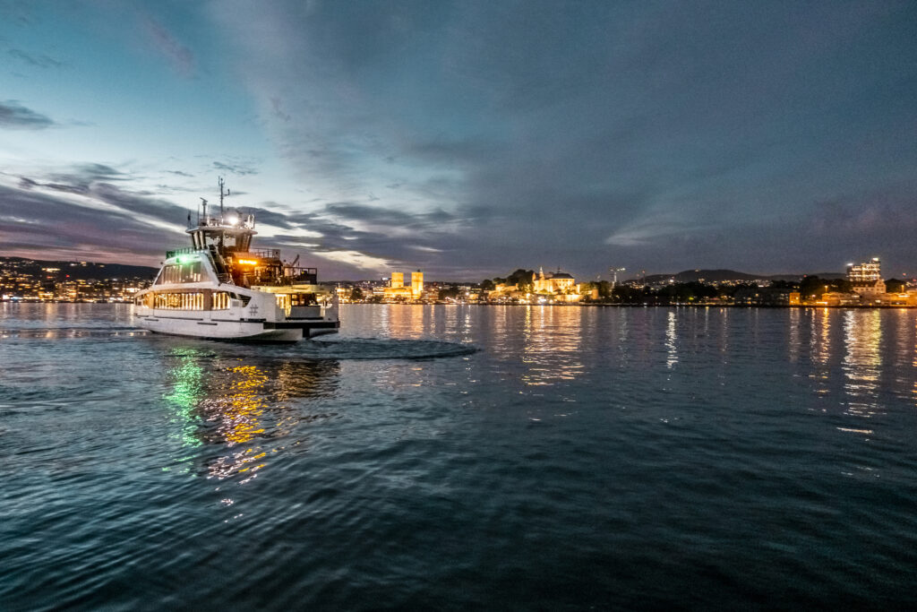 Electrical battery operated ferry at Oslo Fjord at night with illuminated city skyline in the back , 4k resolution