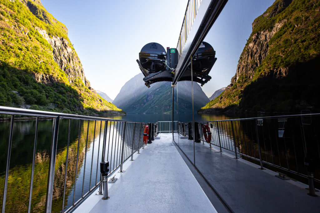 Navigation in the Norwegian fjords have been a vital means of transporting goods and people between communities.  Delivering produce from farms to markets, ferrying private and commercial vehicles and as part of the growing tourist industry.  The infrastructure spans from small private piers and pontoons to large commercial docking stations.  With a comprehensive system of ferry routes, tour boats of various sizes and large cruise liners.  This image was taken in a very calm and bright morning and it shows the reflection of the fjord and mountains on the boat's side window.  The Nærøyfjord is a UNESCO World Heritage Site.