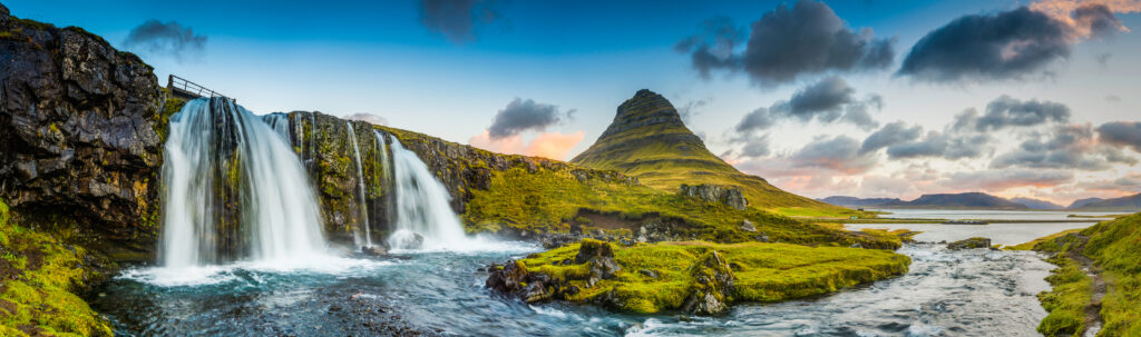 The clear blue waters of Kirkjufellfoss waterfall tumbling down the mountainside to the ocean fjord beside Grundarfjordur beneath the iconic peak of Kirkjufell framed by the warm glow of sunrise over the Snaefellsnes peninsula in the far east of Iceland.