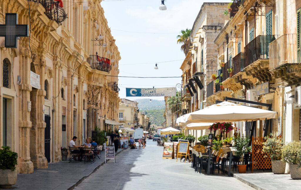 Traditional street in the town of Noto, Sicily, which displays the town's famous Baroque style architecture. Picture taken during a warm summer day and contains some open restaurants and people walking down the street.