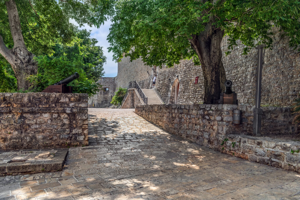 Ancient paved road to the Budva Citadel in the Old Town, Montenegro. Alley with green trees and two ancient cannons among the stone architecture