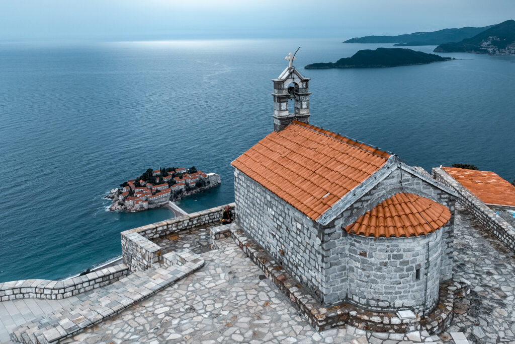 Old church of St. Sava in a small village of Blizikuce captured with a drone and with Sveti Stefan island in a background during one cloudy afternoon in the Adriatic sea.